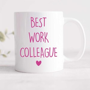 Pode incluir: Caneca de cerâmica branca com uma inscrição rosa que diz "BEST WORK COLLEAGUE" com um coração rosa abaixo do texto.
