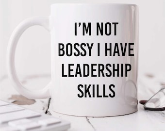 Pode incluir: Caneca de cerâmica branca com o texto "I'M NOT BOSSY I HAVE LEADERSHIP SKILLS" em preto. A caneca tem uma alça curva e está sobre uma superfície branca. Um teclado e óculos são parcialmente visíveis.