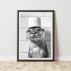 May include: Monochrome framed print of an otter wearing a toilet roll as a hat. The otter is the focal point, set against a white tiled backdrop. The print is displayed on a wooden floor.