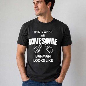 Peut inclure: T-shirt noir avec texte blanc : « THIS IS WHAT AN AWESOME BARMAN LOOKS LIKE ». Le texte est centré, avec deux illustrations de pouces levés en dessous du mot « AWESOME ». Le t-shirt est porté par une personne.