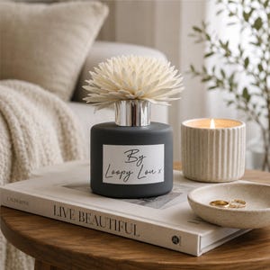 May include: A dark grey diffuser with a white flower-shaped top, a lit candle, and a small bowl with rings on a wooden table. The diffuser has a label that reads "By Loopy Lou x". A book with the words "LIVE BEAUTIFUL" is also on the table.