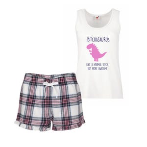 May include: White pajama set featuring a tank top and shorts. The tank top has the text "BITCHASARUS" above a pink dinosaur graphic. The shorts are plaid with white, black, and pink squares and a drawstring.