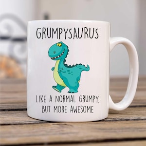 May include: White ceramic mug with the text "GRUMPYSAURUS" above a cartoon dinosaur illustration. The dinosaur is teal with a yellow belly and is walking. Below the illustration is the text "LIKE A NORMAL GRUMPY, BUT MORE AWESOME."