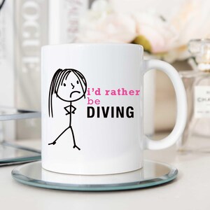 May include: White ceramic mug featuring a black stick figure drawing of a woman with her arms crossed. The text "i'd rather be DIVING" is printed on the mug in black and pink.