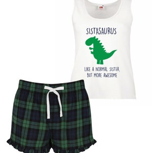 May include: White tank top with a green dinosaur graphic and the text "Sistasaurus Like a normal sister, but more awesome". Green and black plaid pajama shorts with a white drawstring.