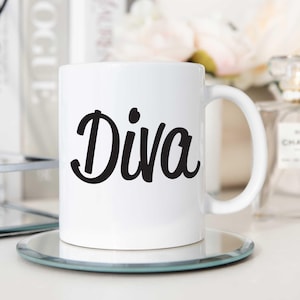 Könnte beinhalten: Weiße Keramiktasse mit dem Wort "Diva" in schwarzer Kursivschrift auf der Vorderseite. Die Tasse steht auf einem silbernen Spiegeluntersetzer.