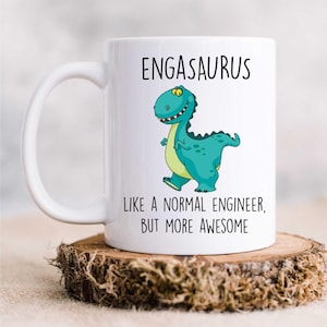 May include: White ceramic mug featuring a green cartoon dinosaur and the text "Engasaurus Like a normal engineer, but more awesome".