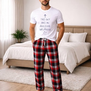 May include: White short-sleeved t-shirt with the text "THIS GUY HAS AN AWESOME GIRLFRIEND" and a smiley face. The shirt is paired with red, black, and white plaid pajama pants with a drawstring waist. The outfit is casual and comfortable.
