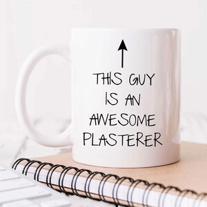 May include: White ceramic coffee mug with a black arrow pointing upwards and the text "This guy is an awesome plasterer".