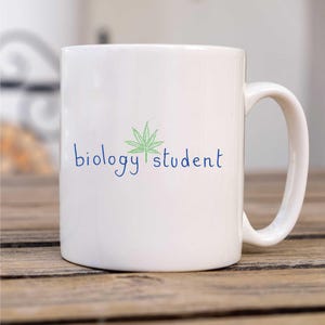 Peut inclure: Une tasse en céramique blanche avec une feuille de marijuana verte et le texte "biology student" en bleu.