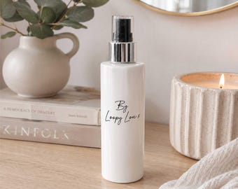 May include: A white spray bottle with a silver and black top. The bottle has the text "By Loopy Lou x" in black script. The bottle is on a wooden surface, with a vase, books, and a candle in the background.