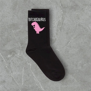 May include: Black socks with the text "BITCHASAURUS" above a pink dinosaur graphic. The socks are made of a soft material and are suitable for everyday wear.
