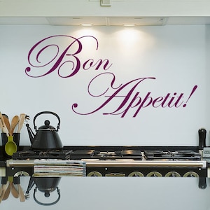 May include: A kitchen scene featuring a black stovetop with a black kettle and utensils. The words "Bon Appetit!" are written in a decorative purple font on the white wall above the stove.