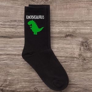 May include: A pair of black socks featuring the white text "KNOBASAURUS" above a green dinosaur illustration. These socks are made from a soft fabric and are designed to fit most adult sizes.