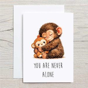 May include: A greeting card featuring a brown monkey hugging a smaller stuffed monkey. The illustration has a soft, watercolor style. The text "YOU ARE NEVER ALONE" is printed in black below the image.