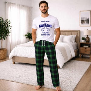 May include: A white t-shirt with blue text reading "THIS IS WHAT AN AWESOME GOLFER LOOKS LIKE". The person is wearing green and black plaid pajama bottoms with a drawstring. The image is taken indoors.