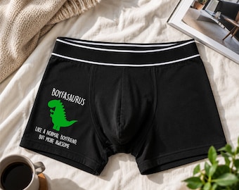 Peut inclure: Boxer noir avec une ceinture blanche et deux rayures blanches. Le boxer présente un motif de dinosaure vert et le texte "BOYFASARUS LIKE A NORMAL BOYFRIEND BUT MORE AWESOME."