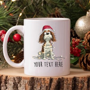 Könnte beinhalten: Weiße Keramiktasse mit einem Cartoon-Hund, der einen Weihnachtsmannhut und Lichterketten trägt. Der Hund ist braun, weiß und hellbraun. Auf der Tasse steht der Text "YOUR TEXT HERE" in Schwarz.