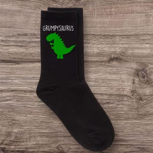 May include: A pair of black socks with a green dinosaur graphic and the text "GRUMPYSAURUS" printed on them.