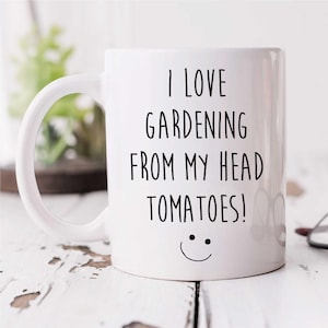 May include: White ceramic mug with the text "I LOVE GARDENING FROM MY HEAD TOMATOES!" in black, and a smiley face. The mug has a curved handle. It is set against a white, weathered wooden surface.