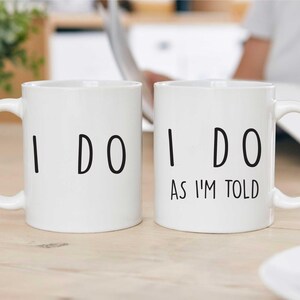 Peut inclure: Deux tasses en céramique blanche avec du texte noir. La tasse de gauche dit "I DO". La tasse de droite dit "I DO AS I'M TOLD".