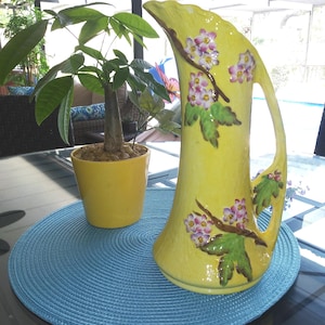May include: A yellow ceramic pitcher with a floral design and a matching yellow potted plant sit on a blue placemat. The pitcher has a curved handle and is decorated with pink and white flowers and green leaves.