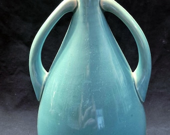 May include: A teal-colored ceramic vase with two curved handles. The vase has a bulbous body that tapers to a narrow neck and flared rim. The handles arch gracefully from the neck to the shoulder of the vase. The surface has a glossy finish.