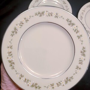 May include: Ivory-colored dinner plates with a floral border. The plates have a silver rim and a delicate pattern of white flowers and green leaves. The plates are round and appear to be made of porcelain.