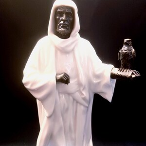 May include: A white ceramic sculpture of a robed figure with a dark face and beard, holding a bronze-colored falcon on his arm. The sculpture has a hood and is set against a black background.