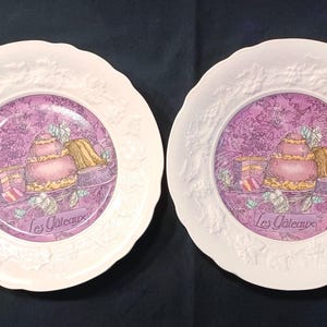May include: Two white decorative plates with a scalloped edge and embossed floral design. The center of each plate features a purple background with a cake illustration and the text "Les Gateaux".