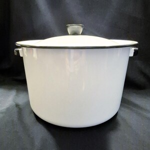 May include: A white enamel pot with a lid. The pot has a black rim and handles. The lid has a black knob. The pot is sitting on a black surface. The pot is a kitchenware item.