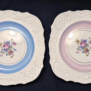 May include: Two square, white decorative plates with a floral design. One plate has a light blue center, the other a pink center. Both plates have a gold rim and a white, textured border.