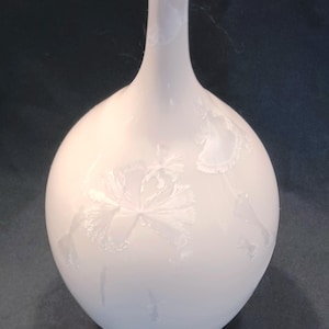 May include: A white ceramic vase with a round body and a long, slender neck. The vase features a delicate floral design etched into the surface. The vase is approximately 8 inches tall.