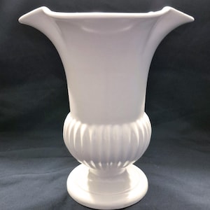 May include: A white ceramic vase with a flared top and a rounded, ribbed midsection. The vase has a wide, circular base and a smooth, glossy finish. The overall design is elegant and classic, suitable for floral arrangements or decorative display.