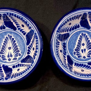 May include: Two matching blue and white ceramic bowls. Each bowl features a central design of a stylized leaf shape surrounded by a circle. The outer rim has a repeating pattern of white leaf-like shapes against a blue background.