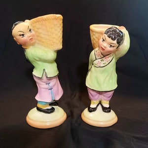 May include: Two ceramic figurines of people wearing traditional Chinese clothing and carrying baskets on their shoulders. The figurines are painted in shades of green, pink, and yellow.