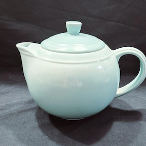 May include: A light blue ceramic teapot with a matching lid and handle. The teapot has a rounded body and a spout for pouring. The lid has a small knob on top. The teapot is designed for brewing tea.