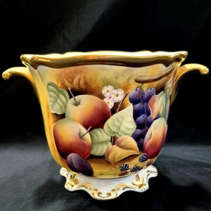May include: A decorative ceramic bowl with a gold rim and handles, featuring a hand-painted design of apples, grapes, and other fruits. The bowl has a white base with gold accents and is set against a dark background.