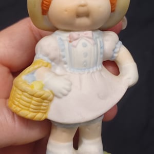 May include: A porcelain figurine of a little girl wearing a white dress, a blue and white patterned bodice, and a straw hat. She is holding a yellow wicker basket and standing on a green base.