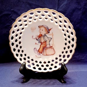 May include: White decorative plate with a scalloped, openwork border and gold trim. The plate features a painted illustration of a young boy with an umbrella and suitcase. The plate is displayed on a black stand.