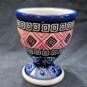 May include: A ceramic egg cup with a blue rim and base, featuring a geometric pattern in pink, black, and white. The cup has a pedestal base and a detailed design.