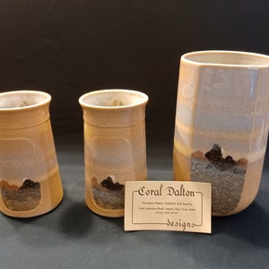 May include: Three ceramic mugs with a brown glaze and a mountain scene design. The mugs are different sizes and the largest mug is taller than the other two. The mugs are on a black surface with a business card that reads "Coral Dalton Designs".