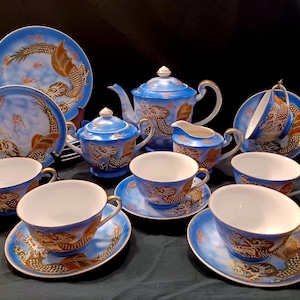 May include: A complete blue and gold porcelain tea set. The set includes a teapot, sugar bowl, creamer, cups, and saucers. Each piece features a detailed dragon design in gold and brown against a blue background. The set is arranged on a dark surface.