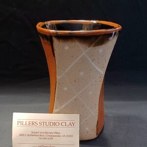 May include: A brown and tan ceramic vase with a white diamond pattern. The vase has a black rim and is slightly curved. The vase is sitting on a white business card with the text "PILLERS STUDIO CLAY" in black.