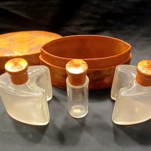 May include: A set of three vintage glass perfume bottles with wooden stoppers and lids. The bottles are in a brown leather box with a decorative pattern.
