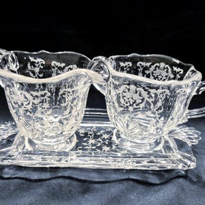 May include: Clear glass creamer and sugar bowl set with floral etched design. The creamer has a handle and spout, and the sugar bowl has a handle. Both sit on a matching rectangular tray with decorative edges.