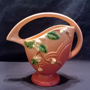 May include: A peach-colored ceramic pitcher with a curved handle and a spout. The pitcher is decorated with white floral accents and green leaves. The base is a darker shade of peach. A decorative piece for the home.
