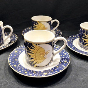 May include: Set of four ceramic coffee cups and saucers. The cups and saucers are decorated with a celestial design featuring a yellow sun and moon on a dark blue background with white stars. The cups have white handles.