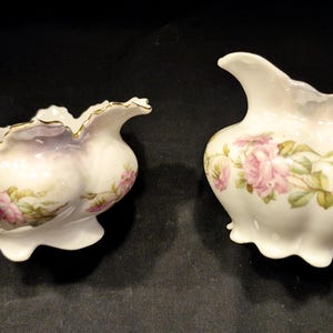 May include: A vintage porcelain creamer and sugar bowl set with a floral design. The creamer has a handle and a spout, while the sugar bowl has a scalloped edge. Both pieces are decorated with pink roses and green leaves.
