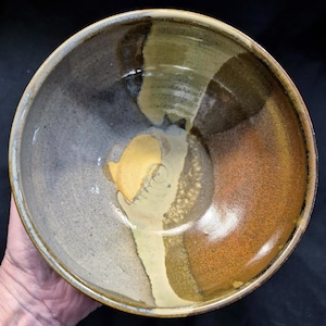 May include: A handmade ceramic bowl with a glossy finish. The bowl features a blend of colors, including gray, brown, and yellow. The interior has a smooth, glazed surface, while the exterior appears textured. This unique bowl is suitable for serving or display.